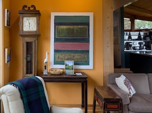 A room with a wooden table, a clock, a painting, a sofa, and a kitchen area. - Tin Roof | Courtyard Retreat Near Bluff Trails (Sea Ranch)