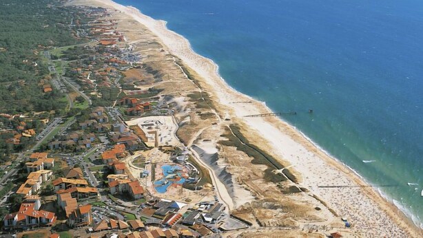 Luftaufnahme einer Küstenlandschaft mit Strand, Wohngebäuden und einem Schwimmbadkomplex.