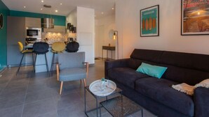 Ein modernes Wohnzimmer mit einem dunklen Sofa, einem gemusterten Sessel und einem Couchtisch. Im Hintergrund ist eine Küchenzeile mit Mikrowelle und Herd zu sehen.