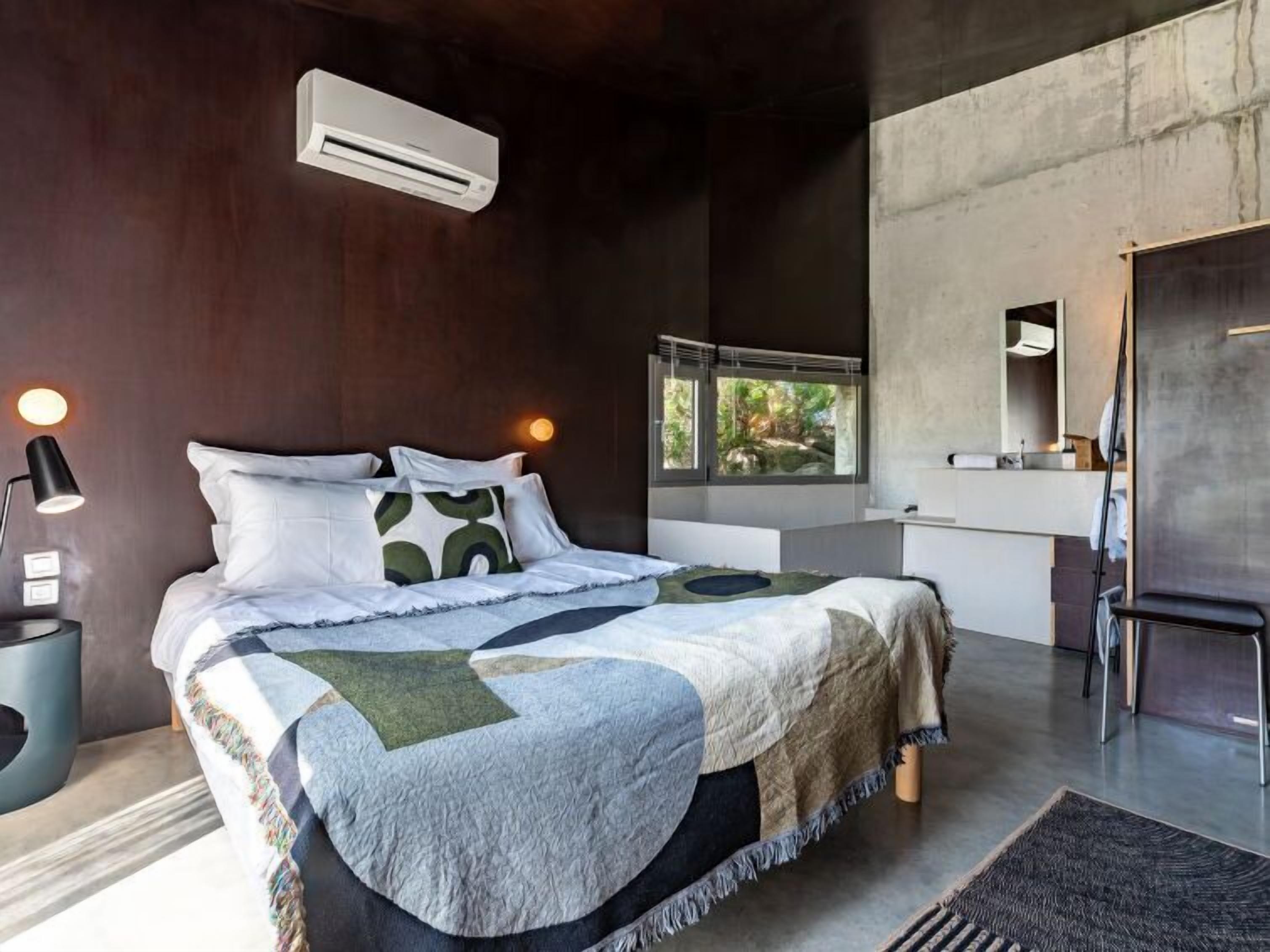 Un dormitorio moderno con una cama grande, una mesita de noche con lámpara, una ventana con vistas a zonas verdes y un equipo de aire acondicionado.