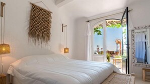 Ein Schlafzimmer mit einem großen Bett, einem gewebten Teppich und Blick ins Freie durch eine offene Tür.