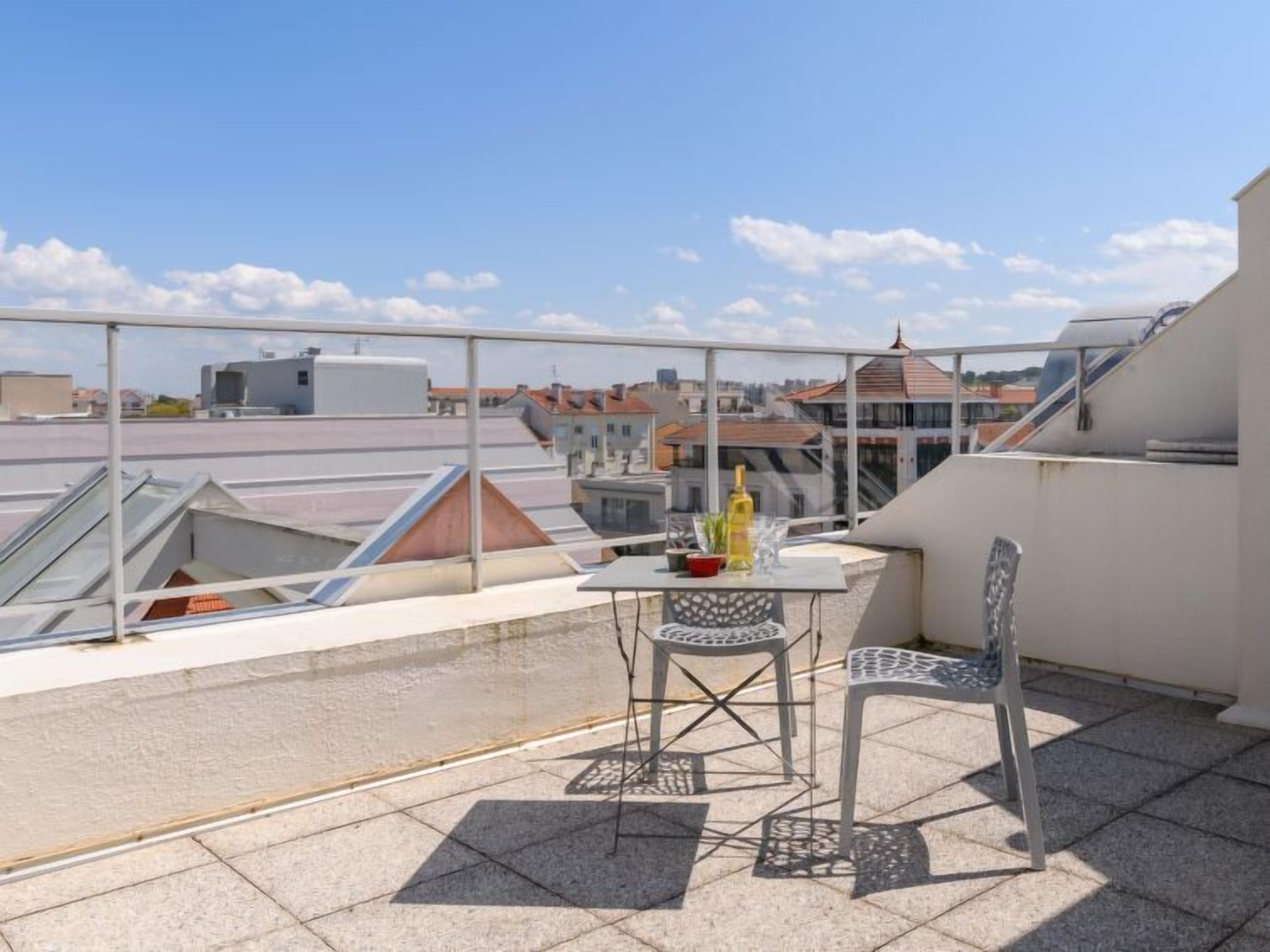 A rooftop terrace with a table and chairs, a glass balustrade, and a view of surrounding buildings.
