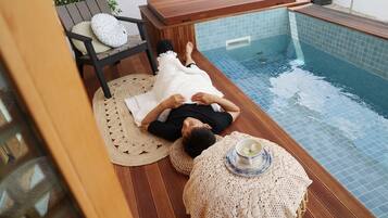 A person relaxing on a wooden deck next to a pool, with a chair and a table with a cup.