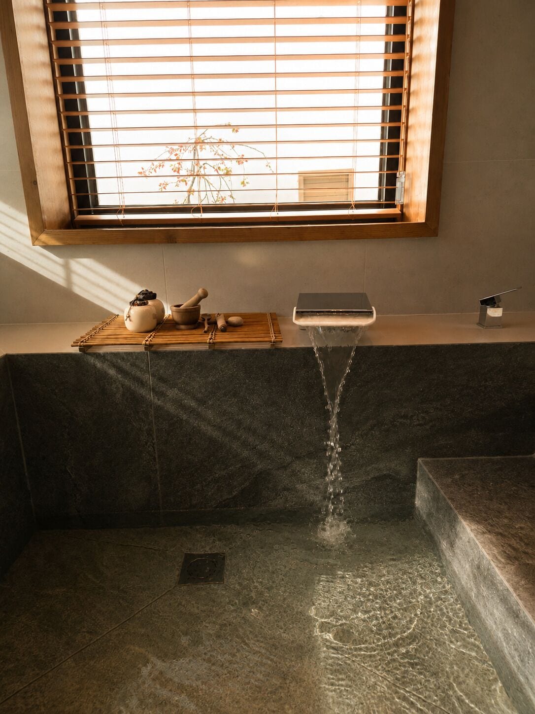 Een badkamer met een badkuip, een raam met jaloezieën, een houten plankje met een theeservies, en een waterkraan.