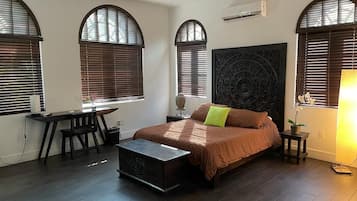 Une chambre avec un lit, un bureau et une chaise. Il y a trois fenêtres avec des stores, un ventilateur de plafond et un appareil de climatisation.