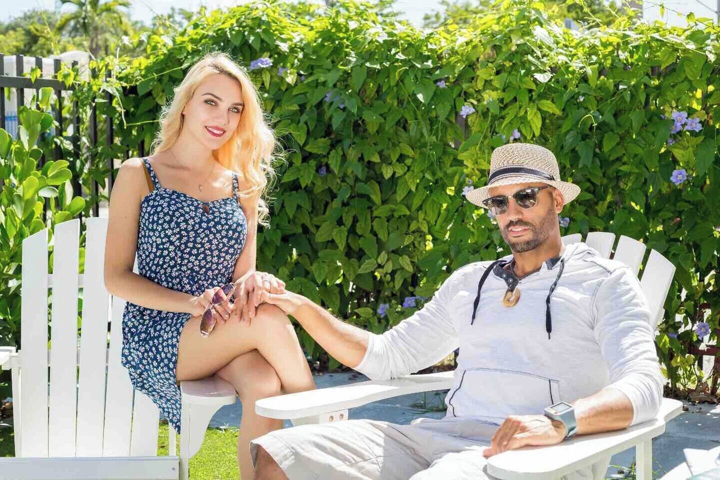 Two people seated on white chairs outdoors with greenery in the background.