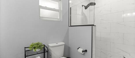 A bathroom with a toilet, a shower with a glass door, a towel rack with a plant, and a window.