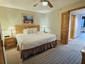 Habitación de hotel con cama, dos mesitas de noche, ventilador de techo, un cuadro en la pared y una puerta que da a otra habitación.