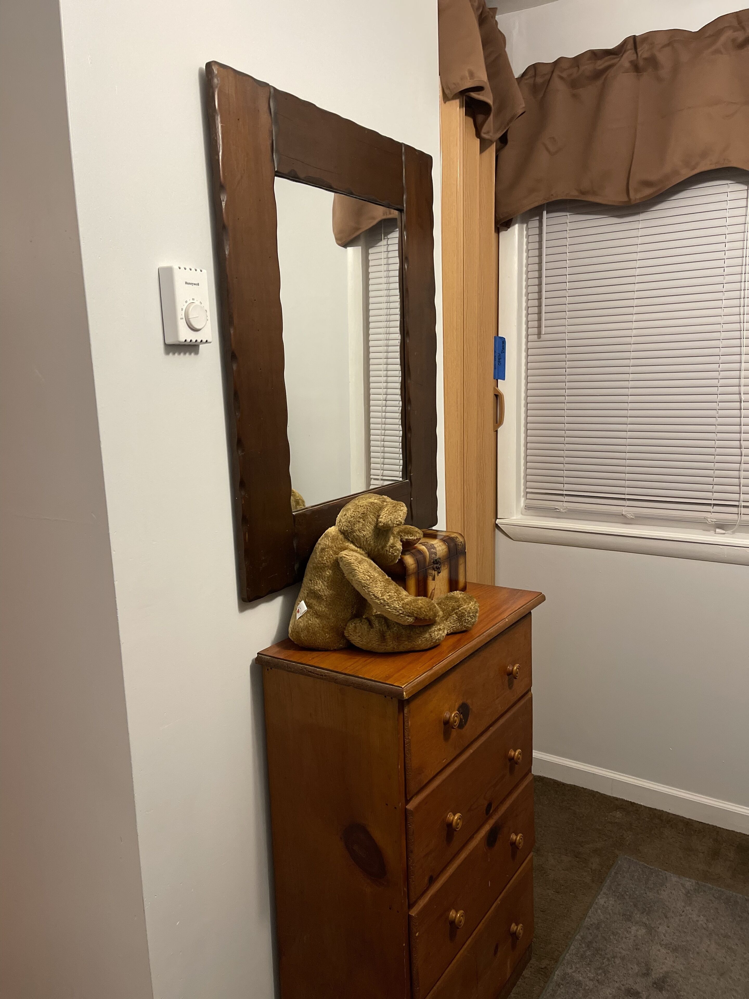 A wooden dresser with a mirror, a teddy bear, and a window with blinds.