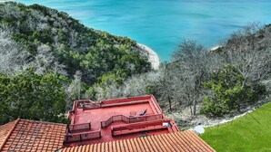 Un edificio de techo rojo con terraza que da a un mar turquesa y una ladera boscosa.