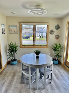 Un coin repas comprenant une table ronde, quatre chaises, deux plantes en pot, un luminaire au plafond et une fenêtre avec des stores.