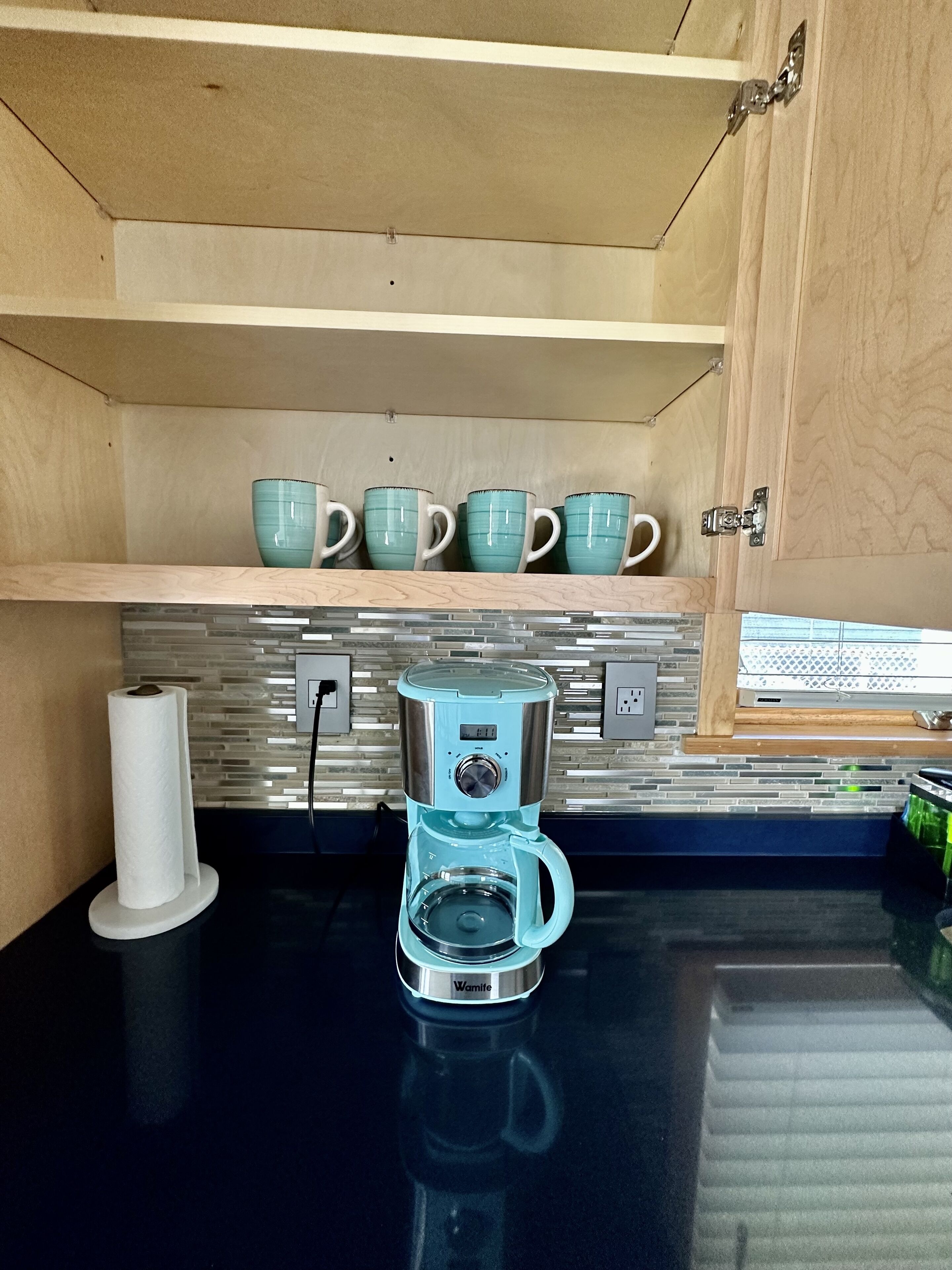 A kitchen with a coffee maker, a roll of paper towels, and a shelf with teal mugs.