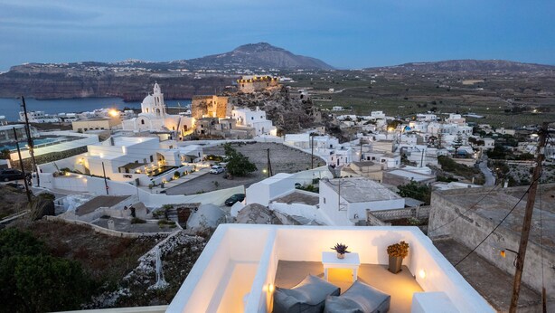 A rooftop terrace with a seating area, white buildings, and a distant view of a fortified structure and coastline. - Santorita Suites (Santorini)