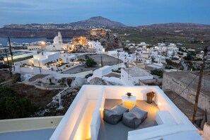 A rooftop terrace with a seating area, white buildings, and a distant view of a fortified structure and coastline. - Santorita Suites (Santorini)