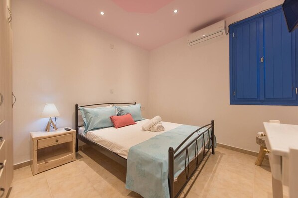 A bedroom with a bed, bedside table, lamp, and a blue wardrobe. - Santorita Suites (Santorini)