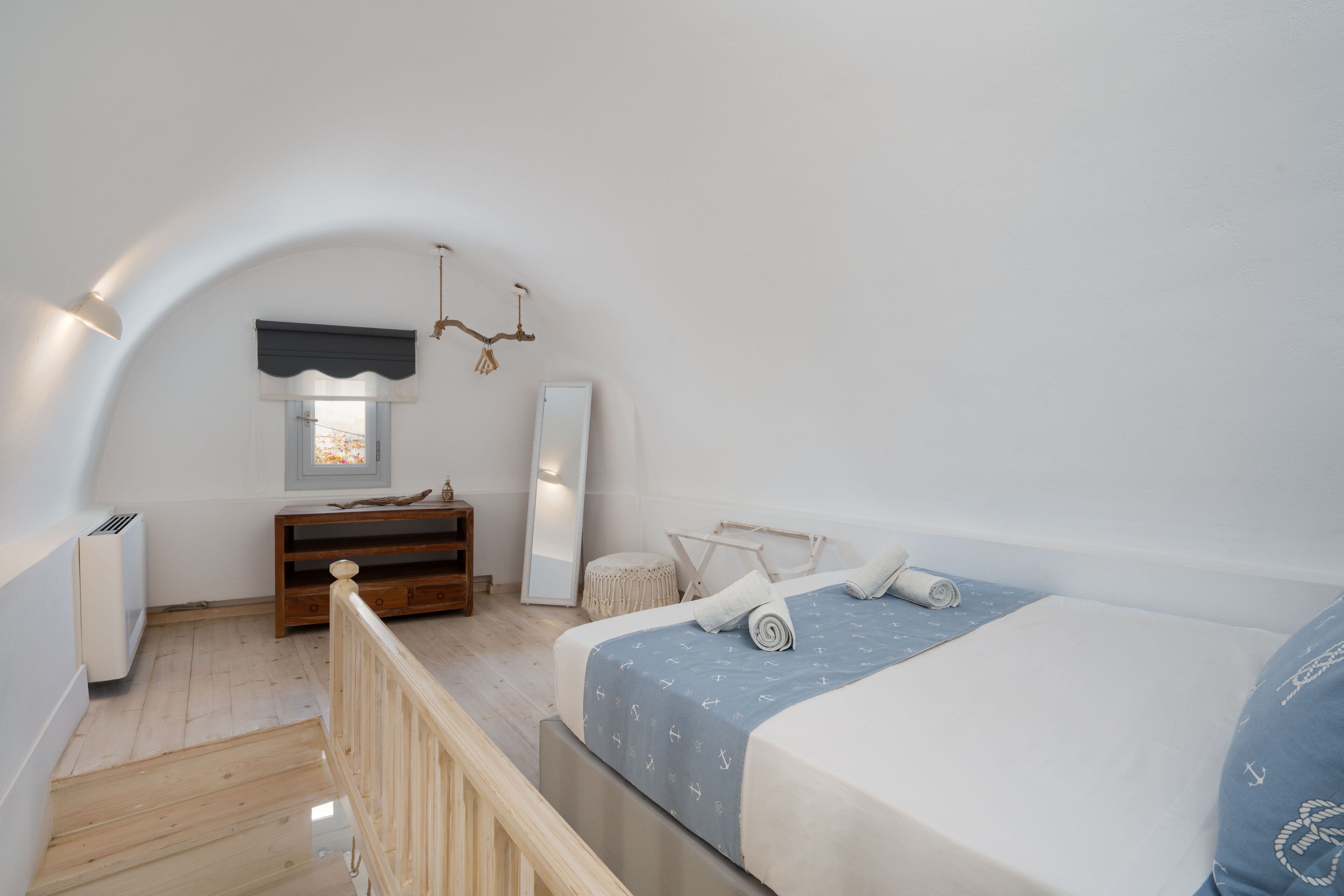 A bedroom with a sloped ceiling, a bed with a blue and white nautical-themed bedspread, a wooden dresser, a mirror, and a small window with a white curtain.