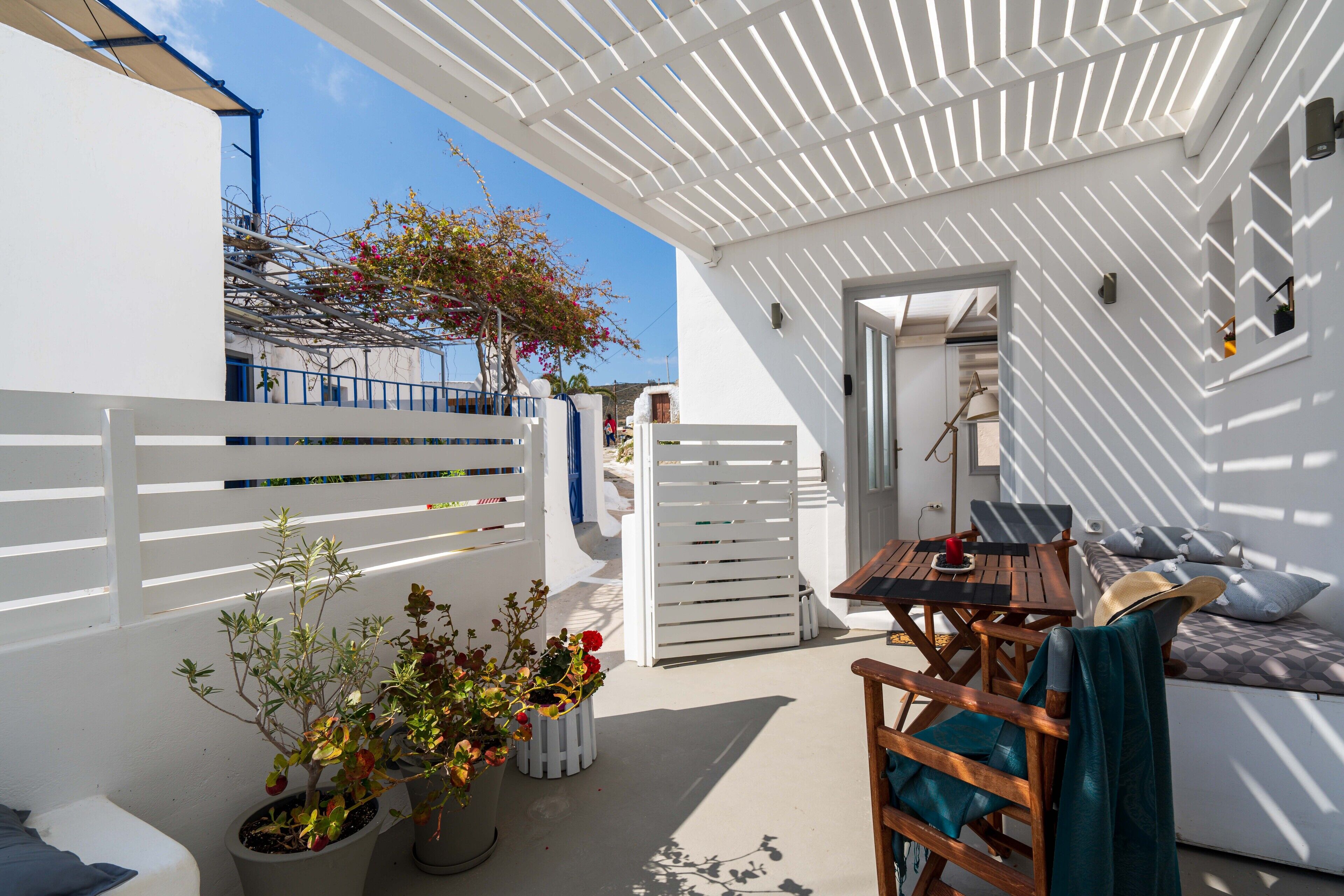 A white-walled outdoor area with a wooden table and chairs, a sofa, and potted plants.