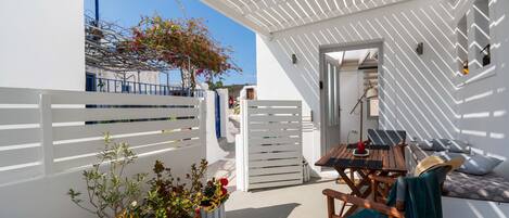 A white-walled outdoor area with a wooden table and chairs, a sofa, and potted plants.