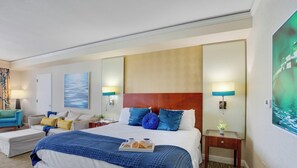 Une chambre d’hôtel avec un grand lit, un coin salon avec un canapé et un fauteuil, un tableau au mur et une fenêtre donnant sur un beau paysage.