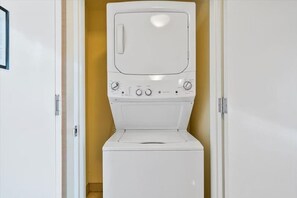 Une machine à laver blanche se trouve dans une pièce avec une porte à gauche et un mur à droite.