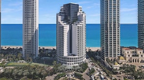 A high-rise hotel with a curved facade, located near a beach with palm trees and a clear blue sky.