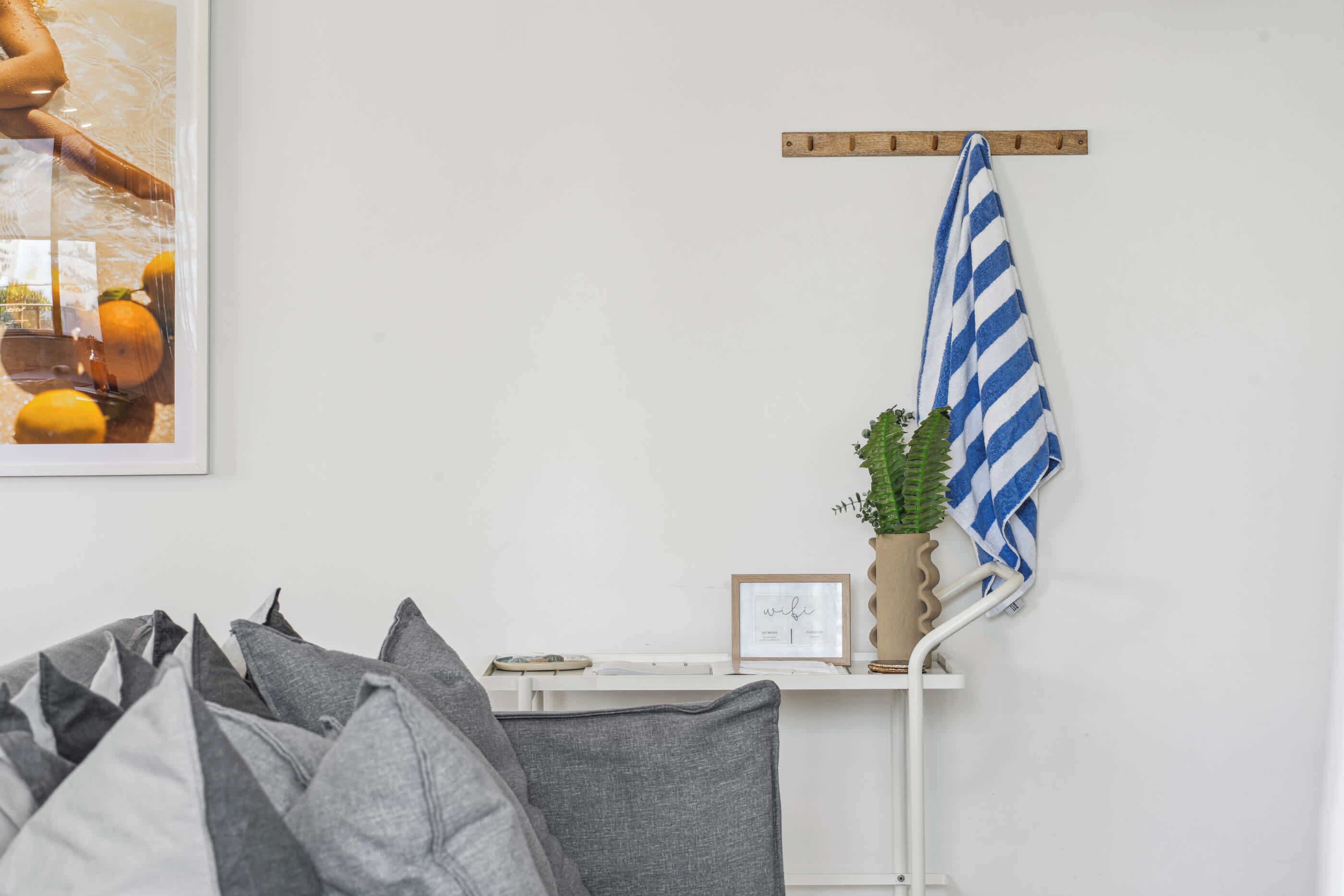 Une pièce avec un mur blanc, une étagère en bois sur laquelle est posée une photo encadrée, une serviette rayée bleue et blanche, et des coussins gris.