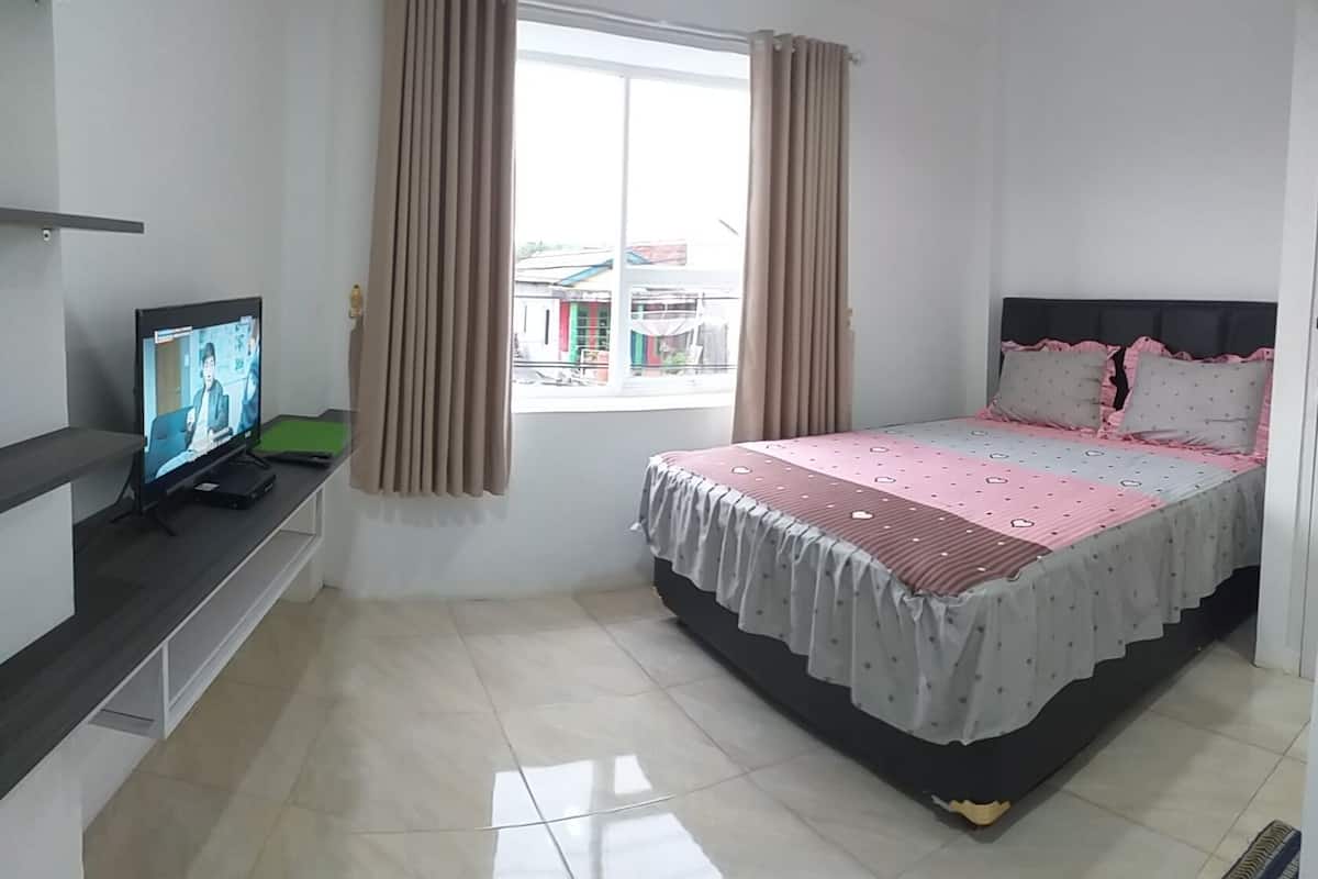 Un dormitorio con cama, televisor sobre un mueble, ventana con cortinas y vistas a edificios.