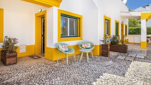 Um pátio com piso de pedra, paredes amarelas e brancas, e duas cadeiras verdes com almofadas coloridas.