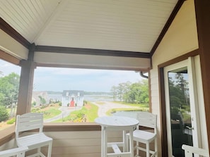 A covered balcony with two white chairs and a small table, overlooking a house and a body of water.