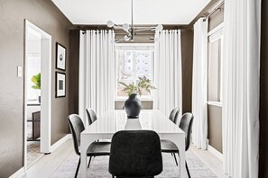 A dining area with a long table, chairs, a vase with plants, and a window with curtains.