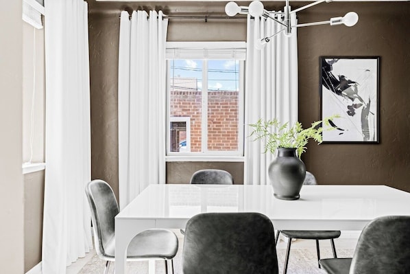 A dining area with a white table, grey chairs, a vase with plants, and a framed picture on the wall.