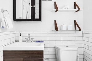 Une salle de bain moderne avec un lavabo blanc, un meuble en bois foncé et des toilettes. On trouve deux étagères en bois au-dessus du lavabo, et une serviette est accrochée au mur.
