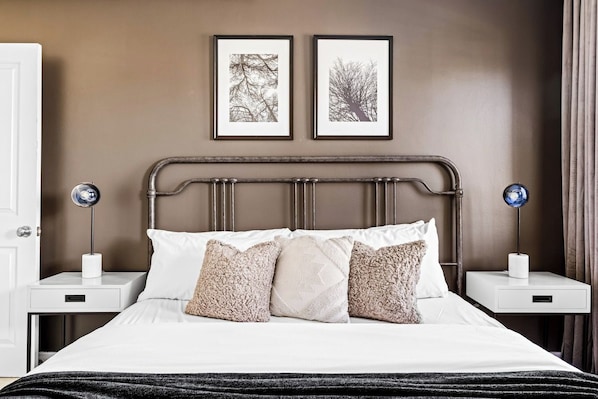 Una cama bien hecha con sábanas blancas y múltiples almohadas, flanqueada por dos mesitas de noche con lámparas, en una habitación con pared marrón y obras de arte enmarcadas.