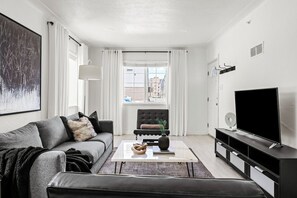 Ein modernes Wohnzimmer mit einer Couch, einem Couchtisch, einem Fernseher und einem Fenster mit Vorhängen.