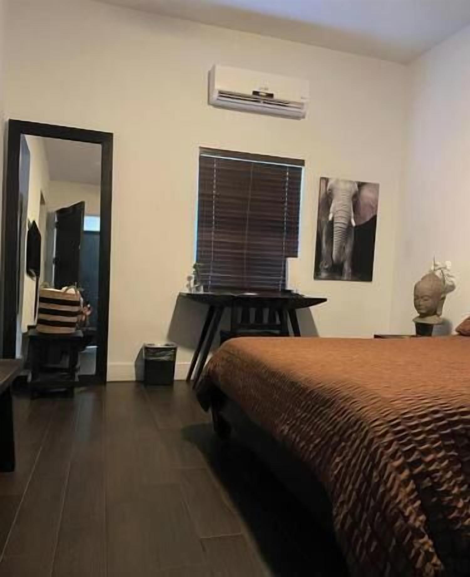 A bedroom with a bed, a desk, a chair, a window with blinds, and a wall-mounted air conditioner.