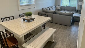 A dining area with a white table and benches, a gray sofa, and a window with blinds. - Sidelines @ Grand Park Game Room, Outdoor Basketball Court Sleeps 13+ (Sheridan)