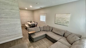 A modern living room with a sectional sofa, a dining table with chairs, and a kitchen area in the background. - Sidelines @ Grand Park Game Room, Outdoor Basketball Court Sleeps 13+ (Sheridan)