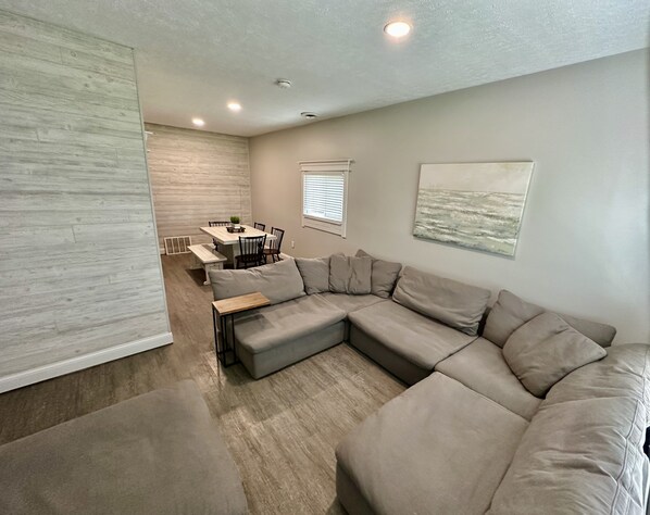 A modern living room with a sectional sofa, a dining table with chairs, and a kitchen area in the background.
