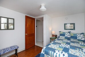 Quarto com porta de madeira, um banquinho, cama com colcha estampada, mesa de cabeceira com abajur e quadro pendurado na parede.