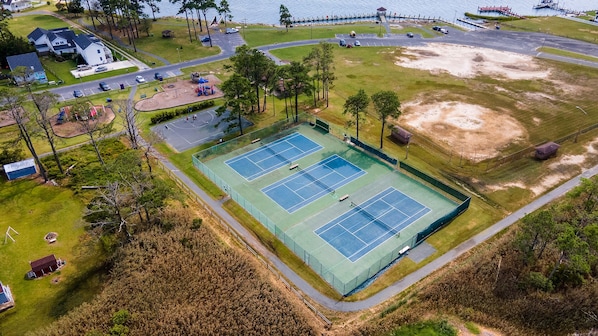 Luftaufnahme eines Tennisplatzkomplexes mit mehreren Plätzen, einem Parkplatz und einem Gewässer im Hintergrund.