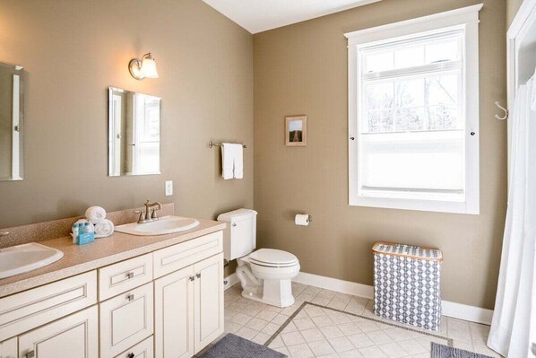 A bathroom with two sinks, a toilet, a trash can, and a window with a view. - Montello Shores (Pets Considered) (West Olive)