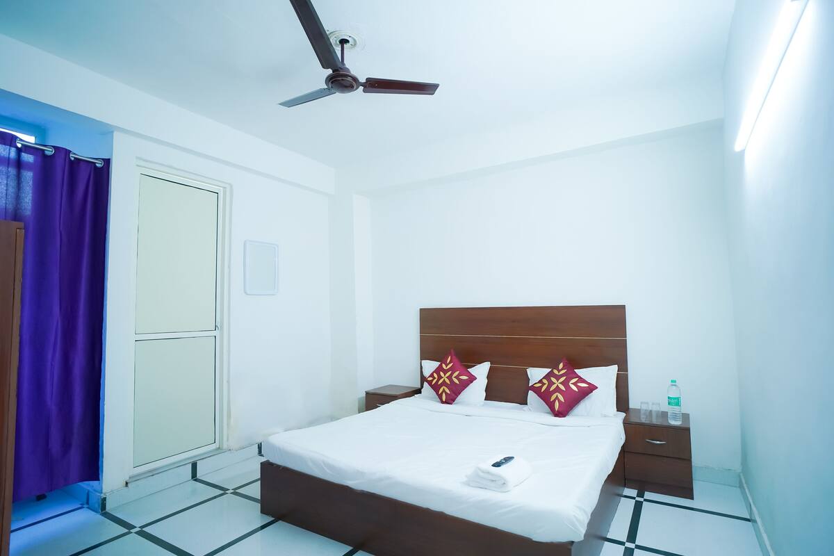 A hotel room with a bed, a nightstand, a ceiling fan, a bathroom door, and a purple curtain.