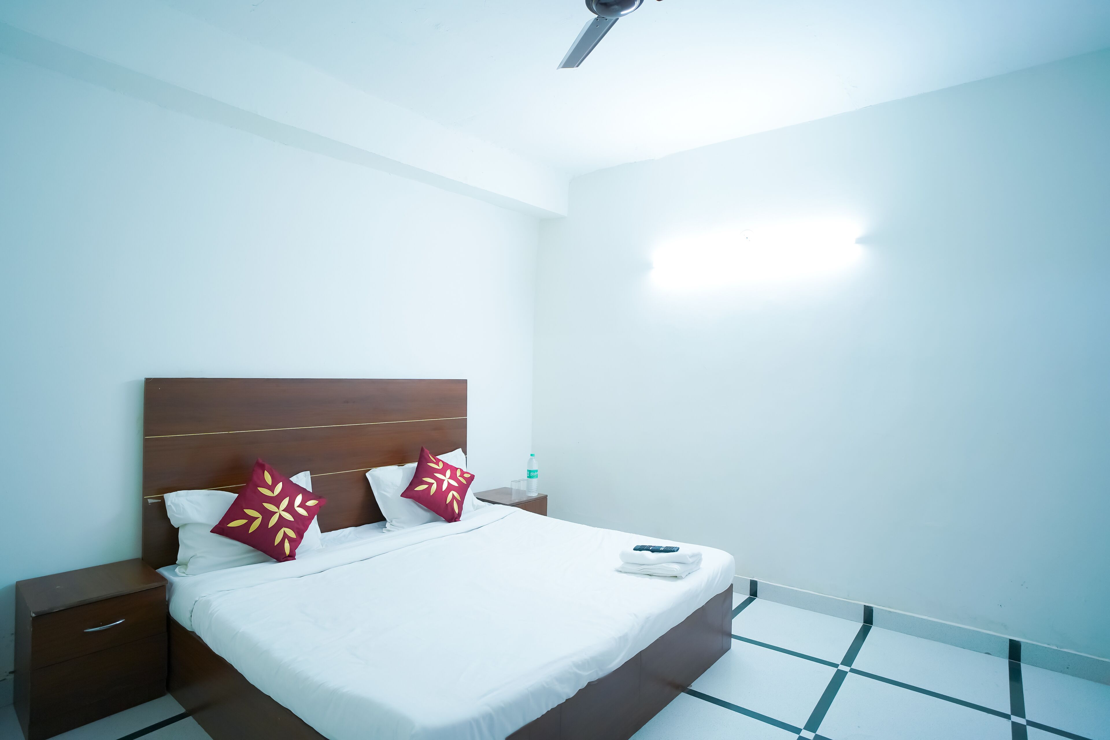 Una cama bien hecha con sábanas blancas y dos almohadones decorativos, un cabecero de madera, una mesita de noche con cajón y una pequeña mesa con una botella de agua.
