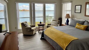 A bedroom with a large bed, a chair, and a small table. There are large windows with a view of a lake. - 3-bedroom home,  waterfront patio,  fishing, kayaking, swimming, yoga room  (Trinity)