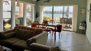 A cozy room with a large window overlooking a lake, a dining area with chairs and a table, and a sofa with a plaid throw. - 3-bedroom home,  waterfront patio,  fishing, kayaking, swimming, yoga room  (Trinity)