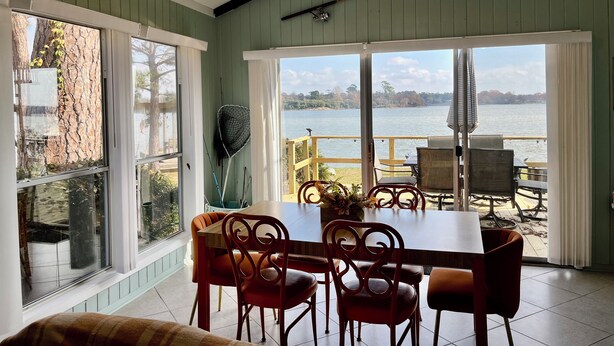 A dining area with a table and chairs, large windows with a view of a lake, and a ceiling fan. - 3-bedroom home,  waterfront patio,  fishing, kayaking, swimming, yoga room  (Trinity)