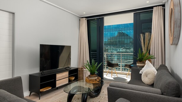 A modern living room with a flat-screen TV, grey sofas, a glass coffee table, and a balcony with a view of the city. - Stylish and Modern Apartment with City Views (Cape Town)