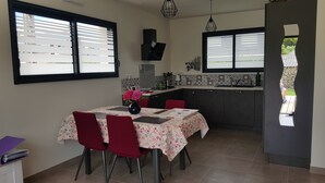 En matplats med ett bord för fyra personer, ett kök med kakel ovanför arbetsbänken och en utsikt genom ett fönster med persienner.