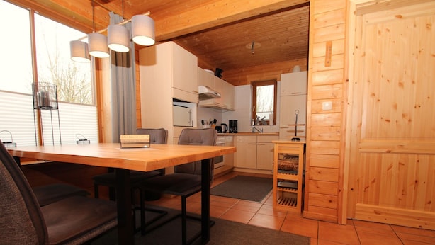 A wooden interior with a dining table, chairs, a kitchen area, and a large window.