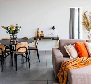 A modern living room with a grey sofa, a dining table with chairs, a small side table with decorative items, and a window with curtains. - Big designed app heart of Brussels (Bruxelles)